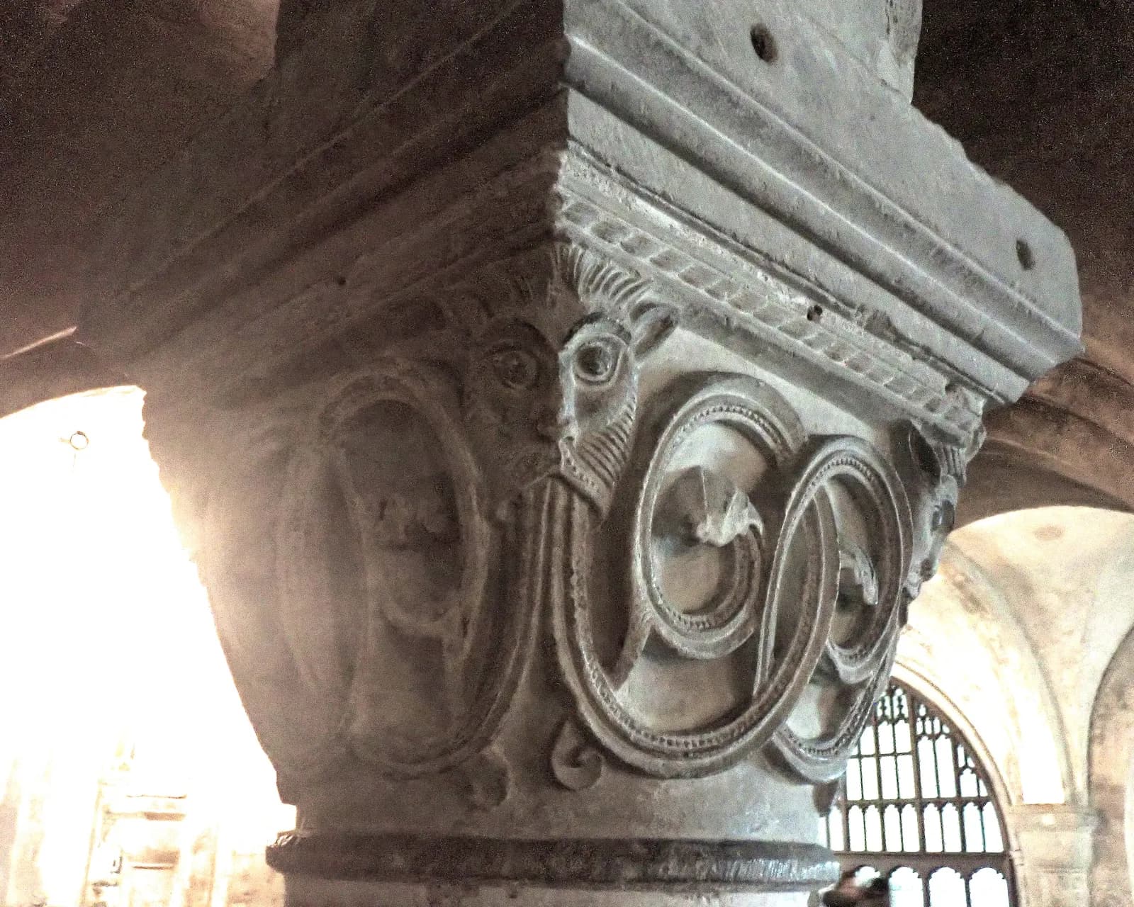 Ornate stone carving on a pillar capital depicting faces emerging from swirling patterns, with light streaming in from the left and an arched window in the background.